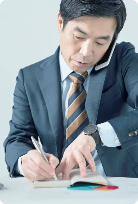 スマートフォンで電話をかけながらメモを取るスーツ姿の男性の画像
